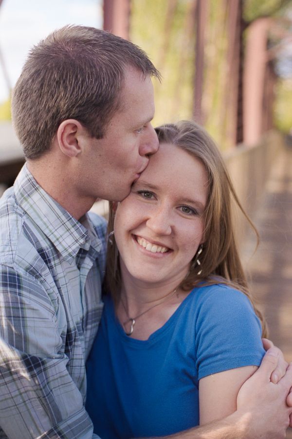 A man in a plaid shirt kisses a smiling woman’s forehead outdoors in a softly lit, natural setting.