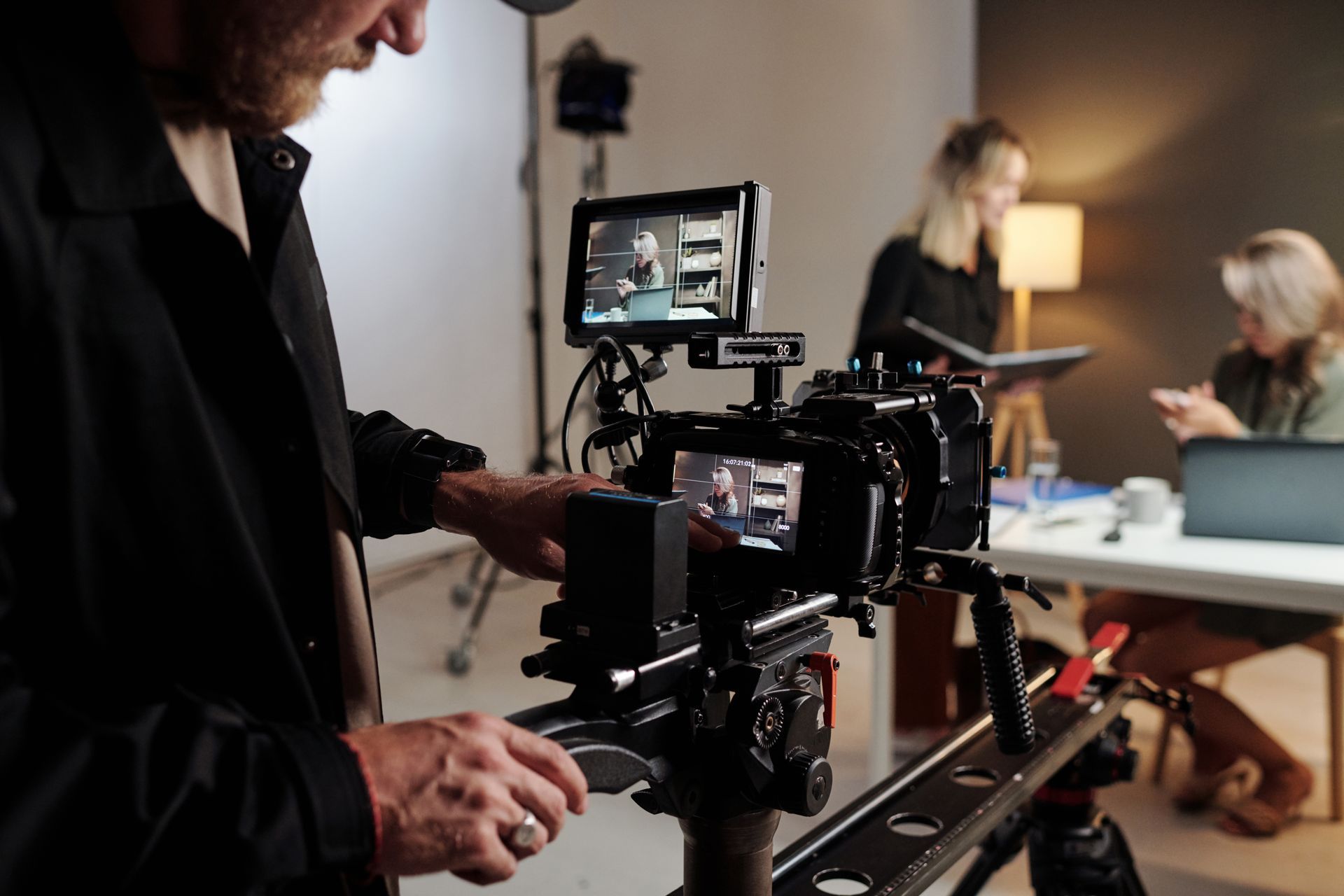 A person operates a video camera on a tripod, filming two people seated at a table.
