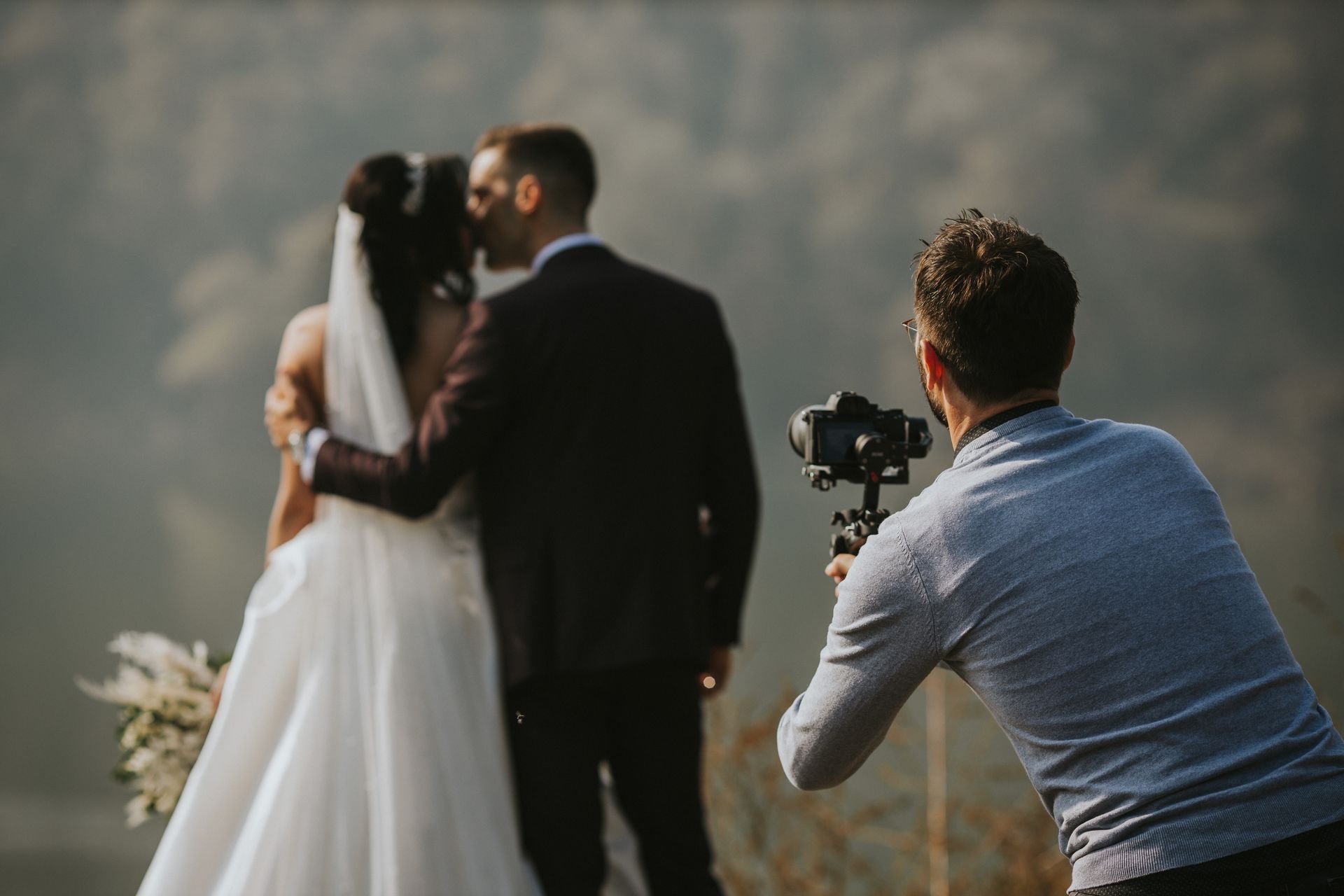 Wedding photographer taking photos of a couple kissing outdoors, blurred background of water and sky.