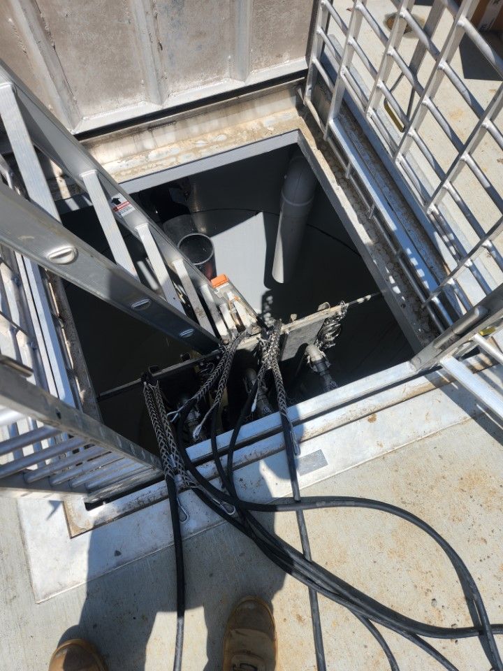 Ladder descending into a dark hatch; metal railing surrounds the opening, black cables, pipes, and equipment visible.