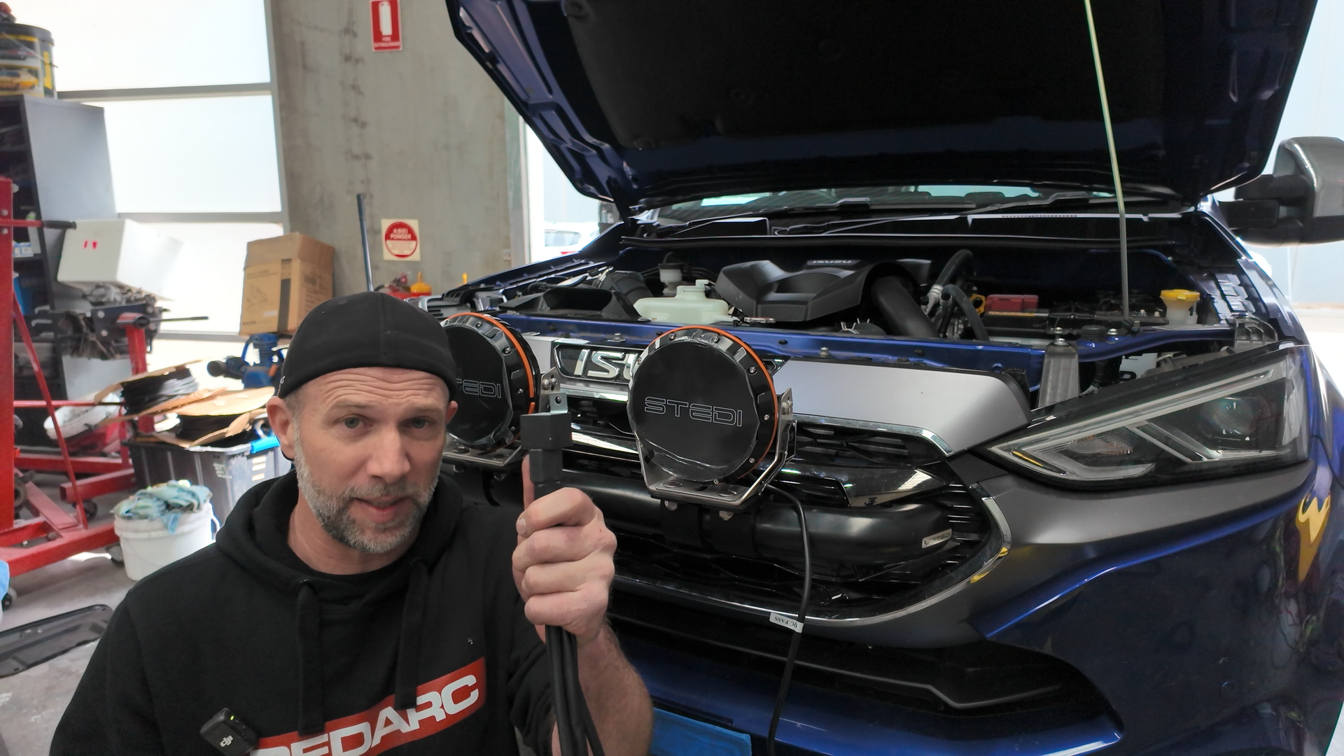 Man holding a cable, standing in front of a blue car with hood open. Workshop setting.