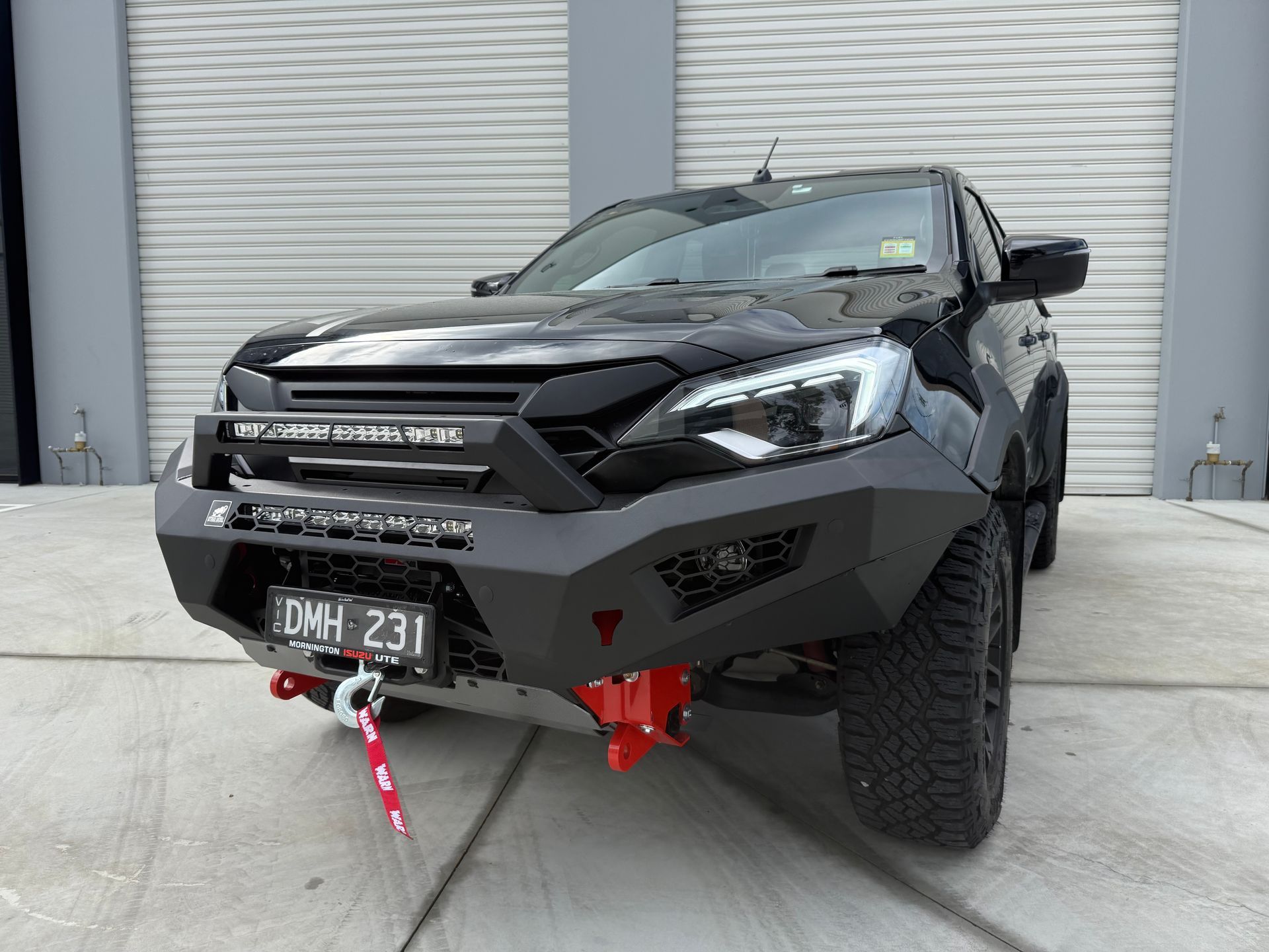 Black pickup truck with custom bumper, winch, and red tow hooks, parked in front of closed garage doors.