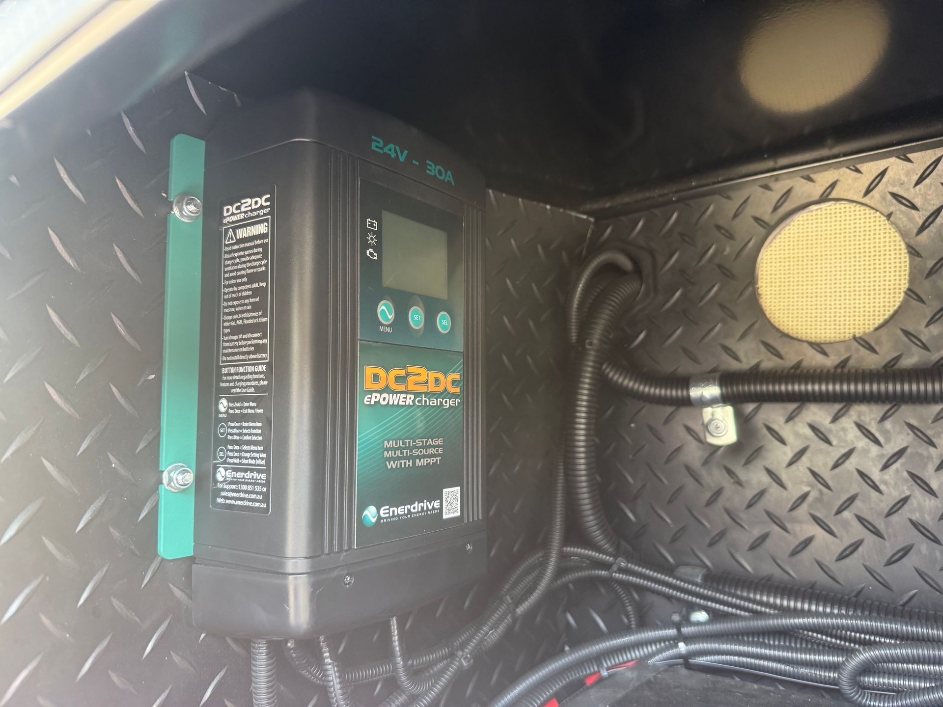 DC-DC battery charger mounted inside a metal compartment. Black and green charger with display screen and wiring.