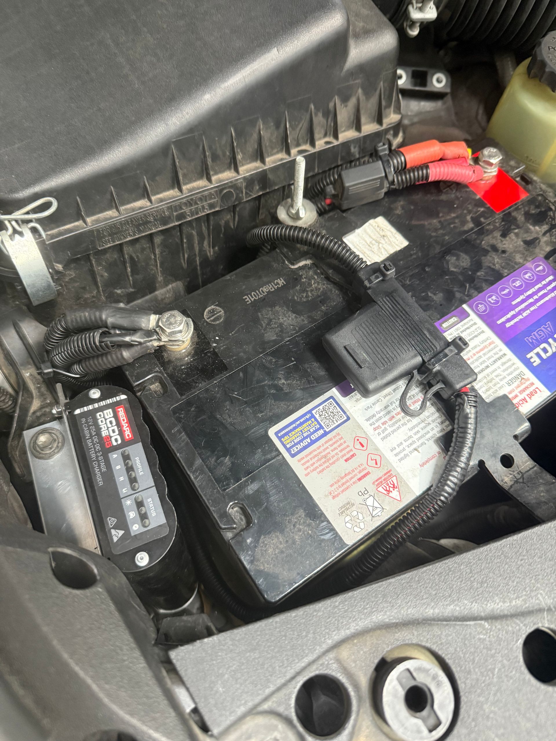 Car battery with attached wiring, fuses, and a black box in the engine bay.