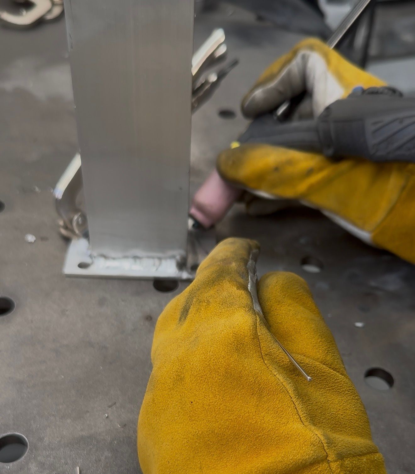 Welder in yellow gloves TIG welding a metal post to a base plate.