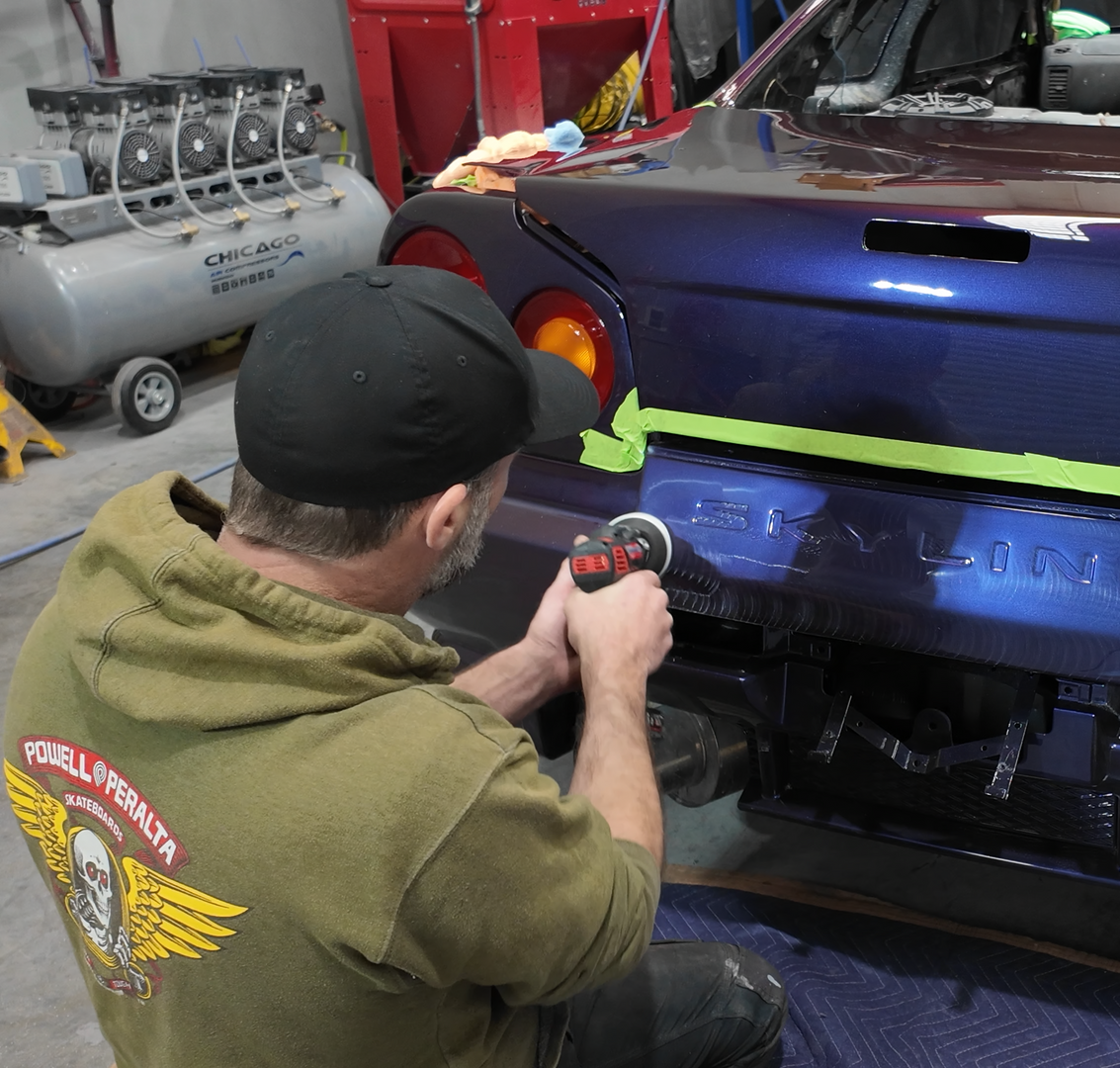 Person sanding a car bumper in a workshop, wearing a green hoodie and black cap.