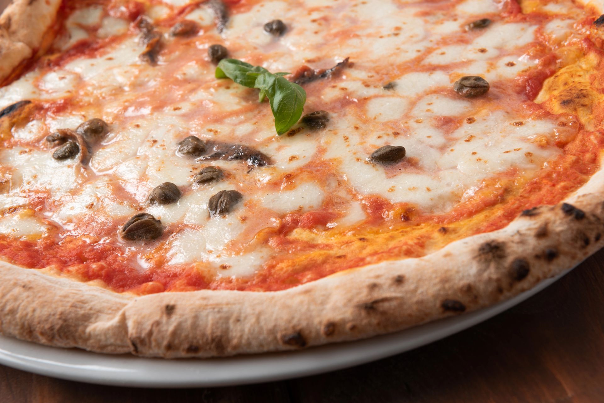 Una pizza intera con salsa di pomodoro, mozzarella fusa, acciughe, capperi e una foglia di basilico fresco su un piatto bianco.