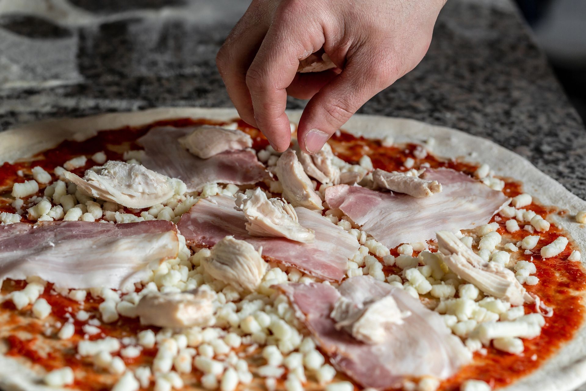 Una mano posiziona dei pezzi di pollo su una pizza condita con salsa di pomodoro, formaggio e sottili fette di prosciutto.