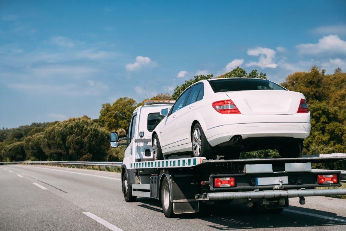 White car being towed on a flatbed tow truck on a highway with green trees and a blue sky.