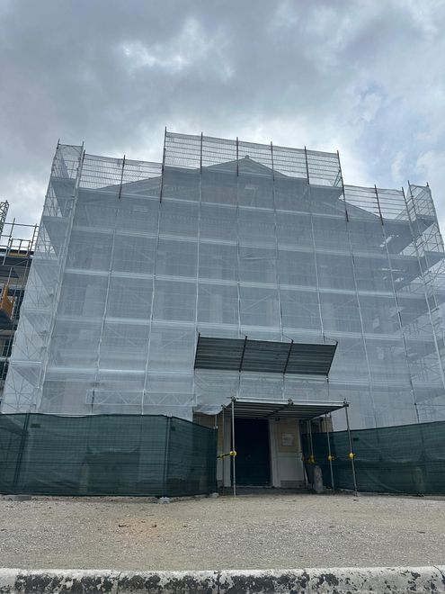 Edificio impalcato e coperto da una rete bianca sotto un cielo nuvoloso, con un primo piano ghiaioso e l’ingresso aperto.