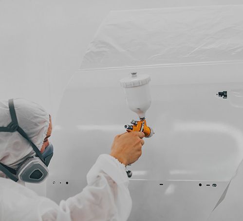 Painter in white protective gear spraying paint on a car body in a clean workshop
