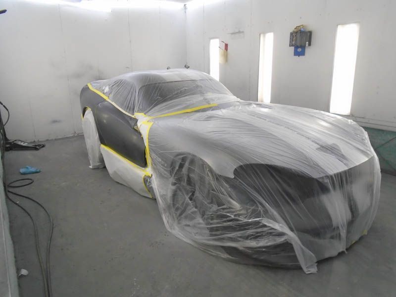 Car covered in plastic sheeting inside a white auto paint booth, masked with tape for painting