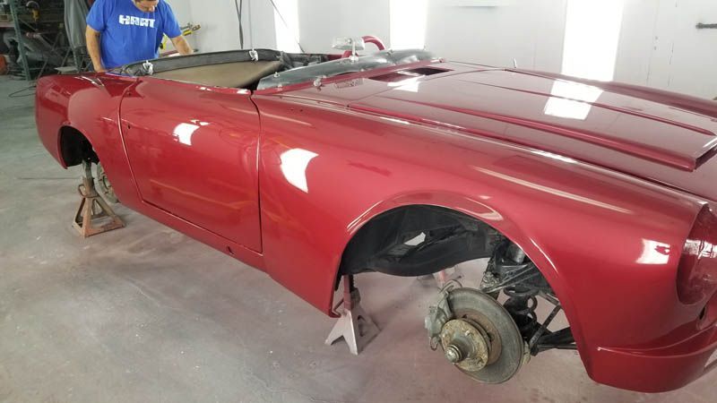 Red car body shell in a garage, missing wheels, with exposed brake assembly and a person standing nearby