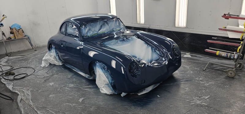 Dark blue classic car body in a paint booth, partly masked with plastic sheets and tape