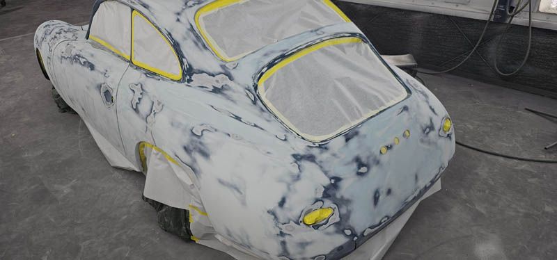 White car body in primer with yellow tape masking windows, parked in a workshop garage.