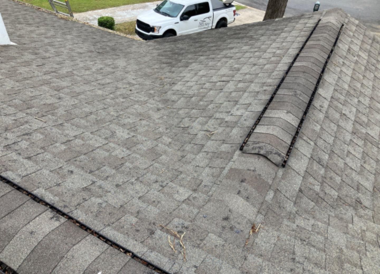 Shingled roof with a raised ridge vent, viewed from above near a parked white truck.