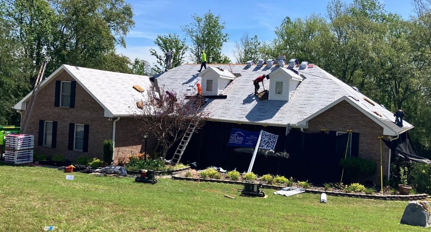 Damaged house with collapsed roof and broken front facade, surrounded by debris and trees in a sunny yard