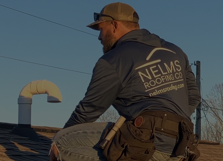 Roofing worker on a rooftop at sunset, wearing a safety harness and branded jacket.