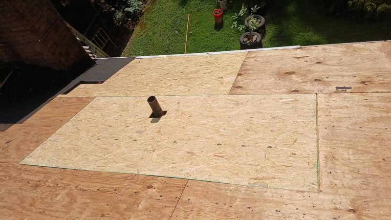 Partially installed plywood roofing with a small vent pipe and green yard in the background