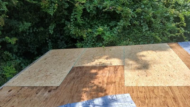 Plywood sheets laid on a roof under construction beside dense green trees.
