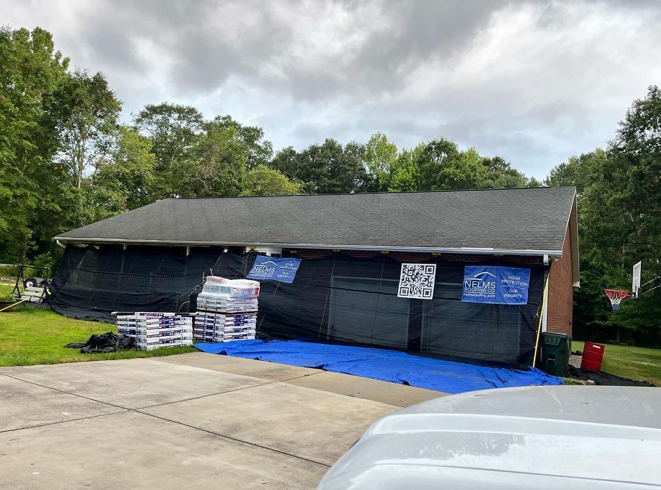 Single-story house with black siding, blue tarp, and signs, viewed from a driveway under cloudy skies