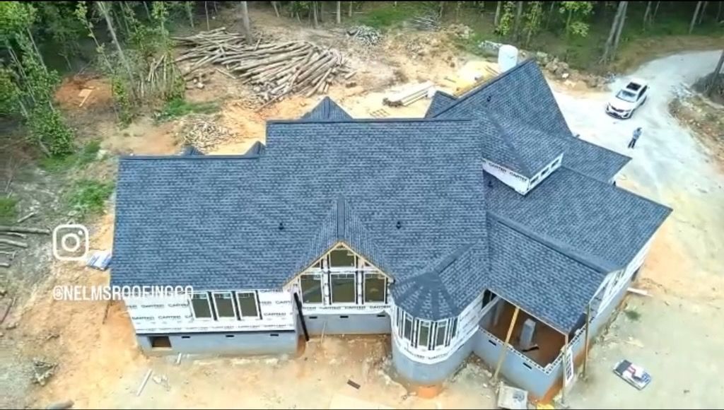 Aerial view of a large dark-blue roofed house under construction on a wooded lot.