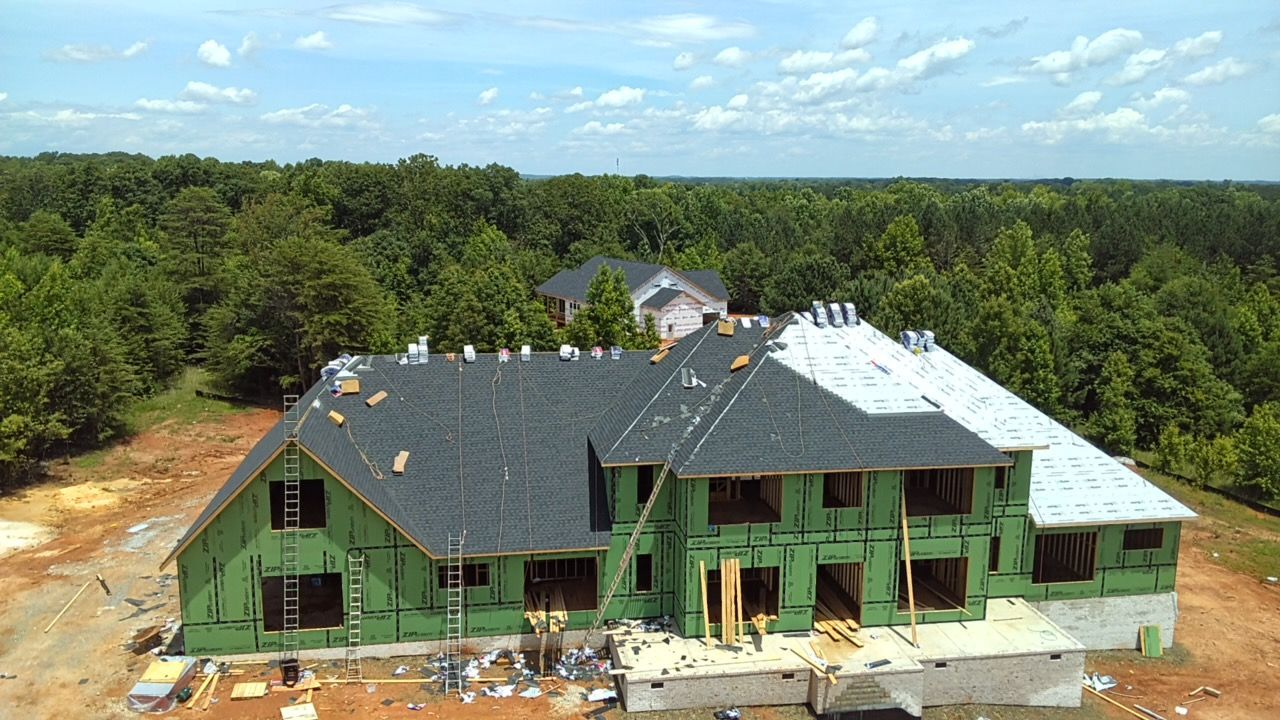Partially built house with green sheathing and white roof in a wooded area under a blue sky
