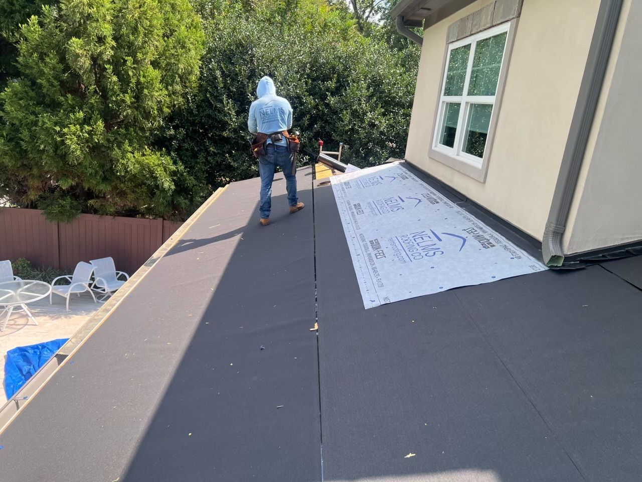 Worker installing roofing on a sloped roof beside a house with trees nearby
