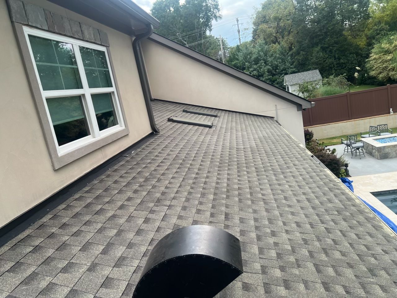 Sloped shingled roof beside beige house with a vent and tree-lined yard in the background