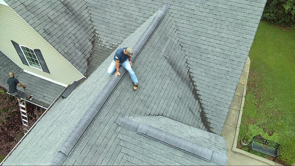 Person sitting on a gray shingled roof near the ridge, with a ladder visible at the left edge.