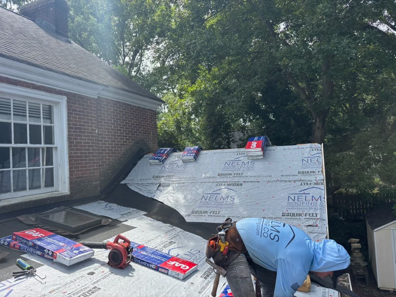 Workers installing white roofing panels beside a brick house, with tools on the roof in daylight.
