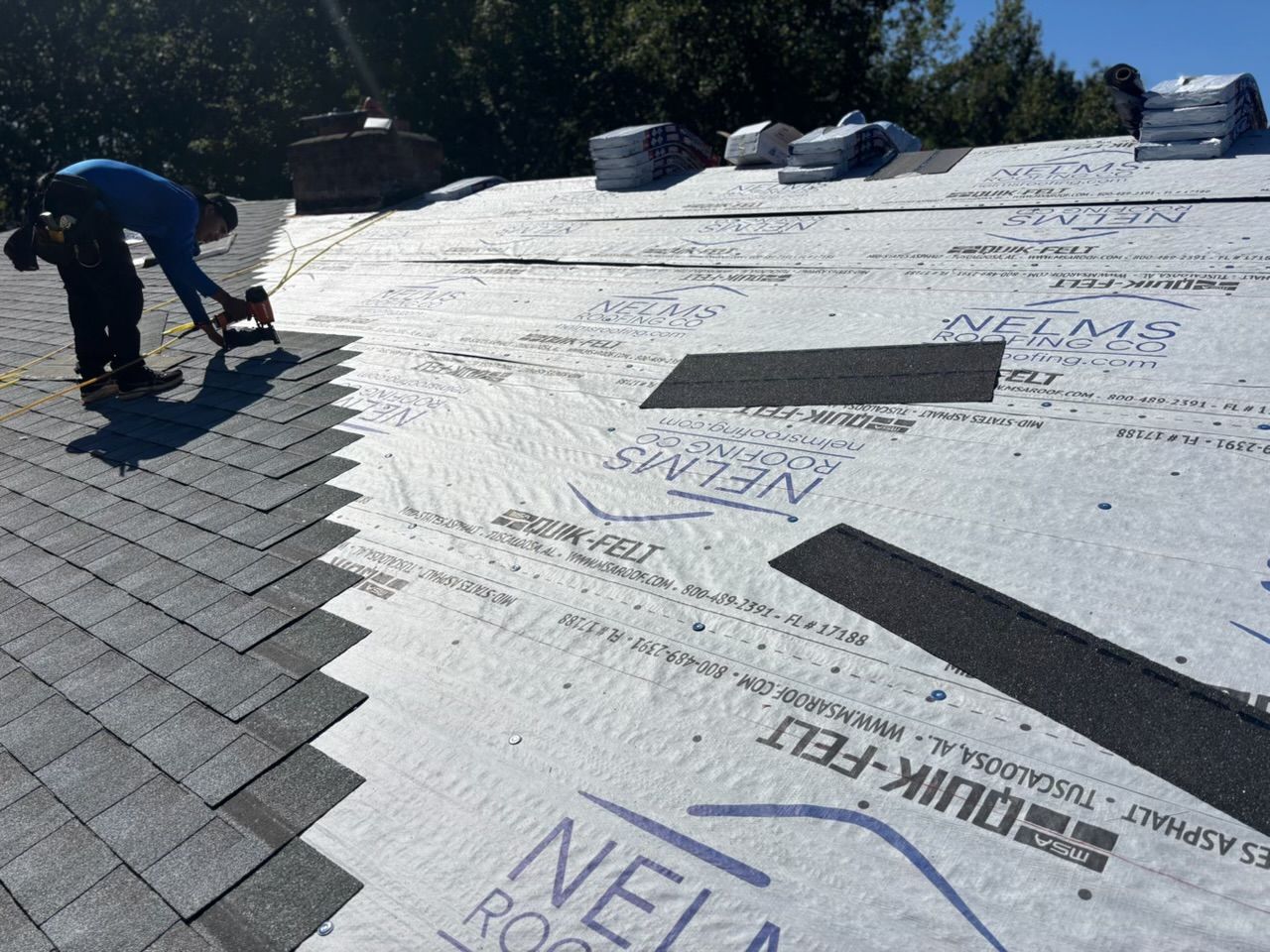 Worker installing underlayment on a sloped roof with gray shingles and white wrap