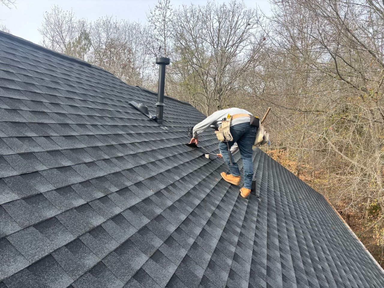 Worker inspecting steep shingled roof near a chimney on a wooded hillside