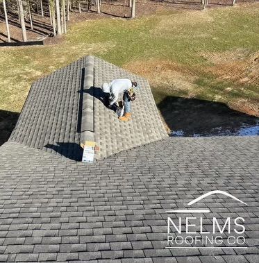 Worker installing shingles on a gray roof above a wooded yard, with NELMS Roofing Co. watermark
