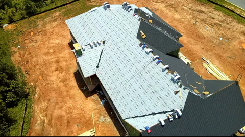 Aerial view of a house under construction with a partially installed roof on a dirt lot.