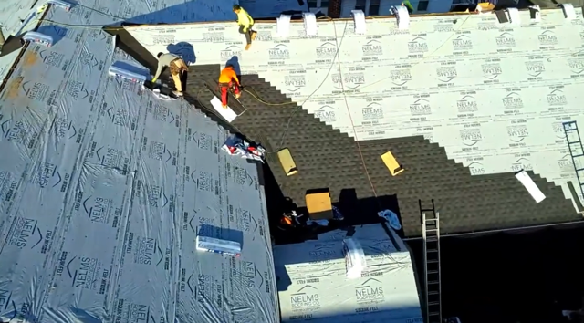 Workers on a roof installing dark shingles beside exposed white roofing underlayment and a ladder.
