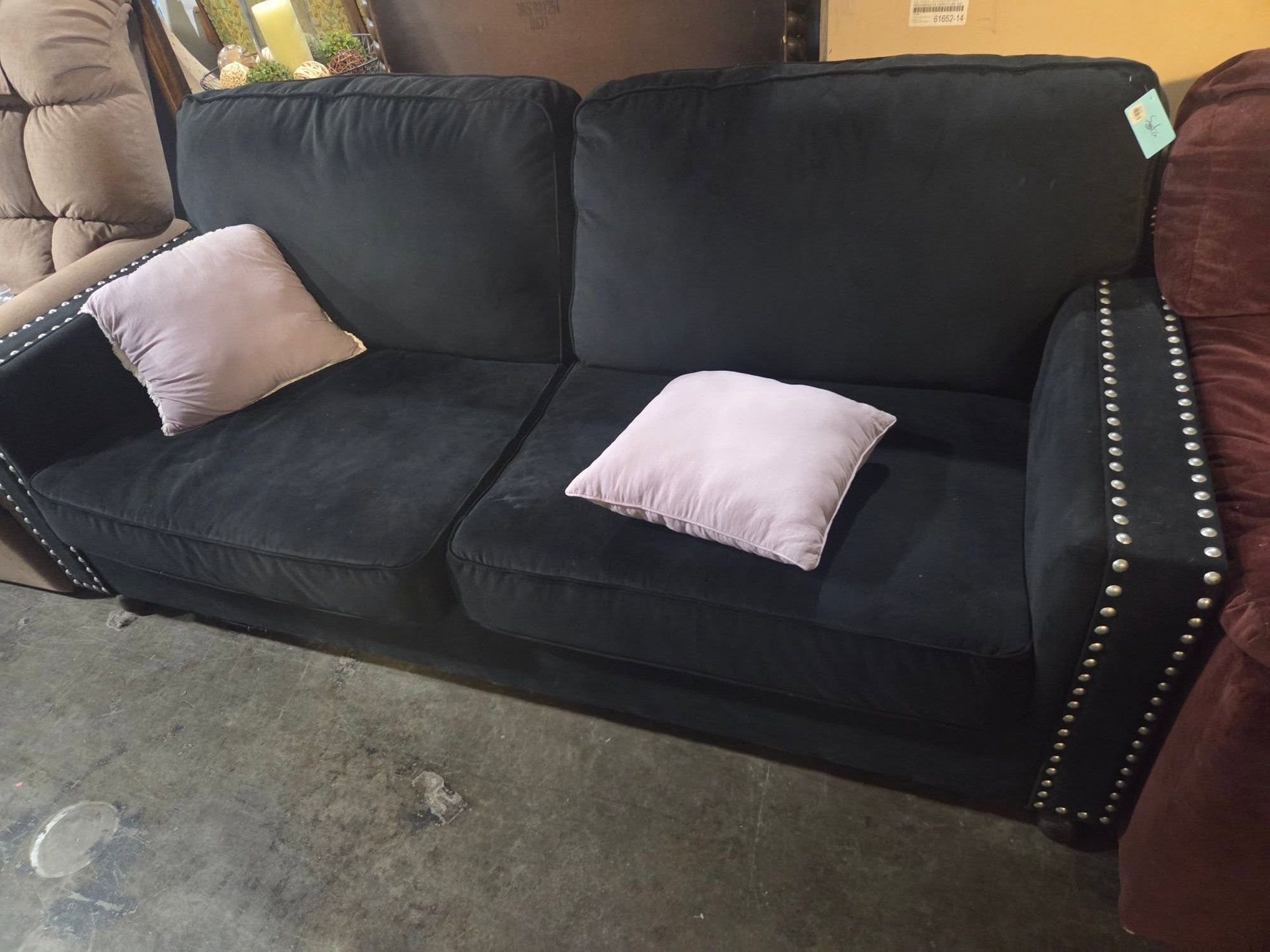 Black fabric sofa with silver nailhead trim and two pink pillows on a concrete floor.