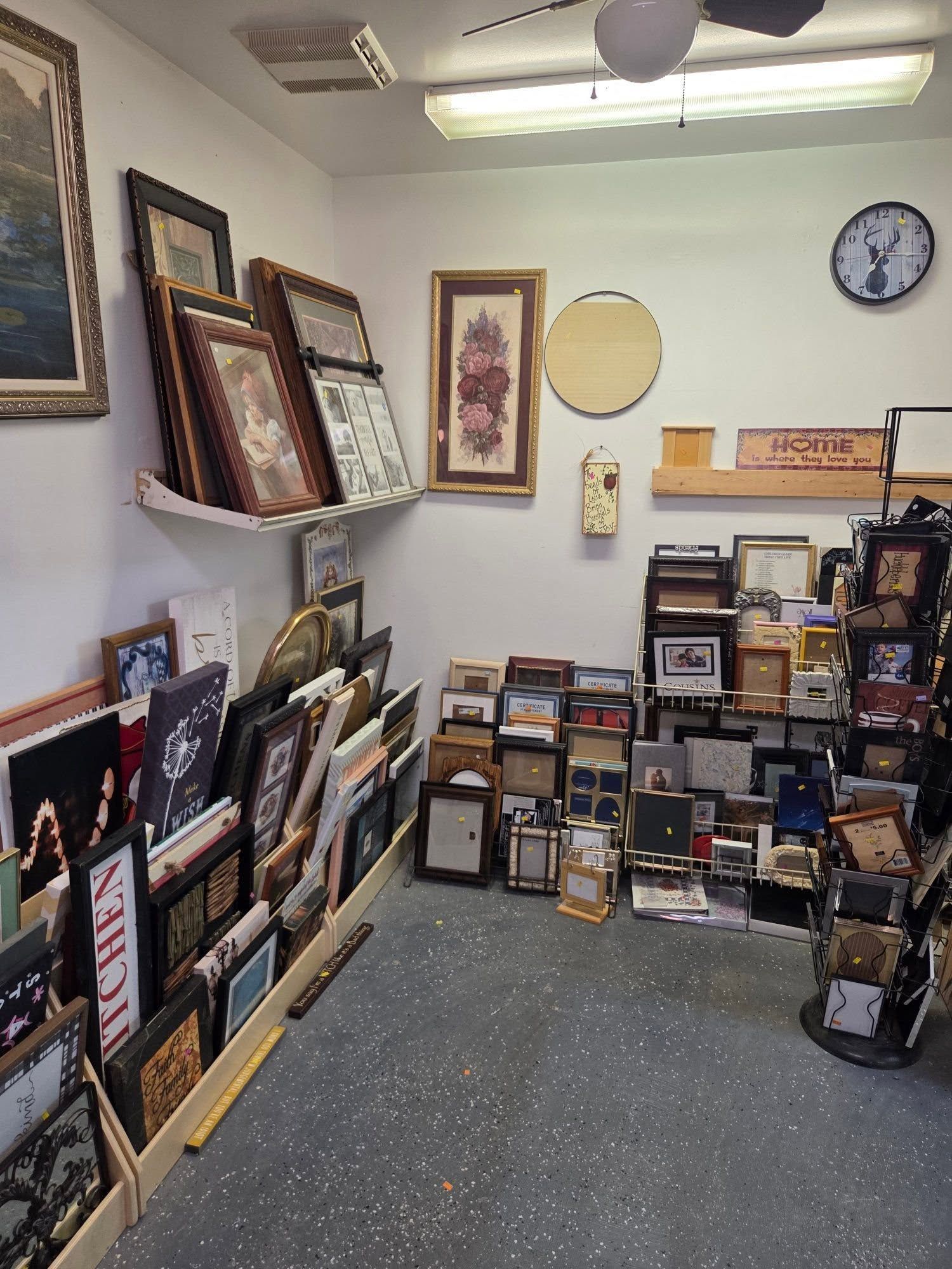 Art shop interior with framed prints and paintings stacked along the walls and floor, clock on the wall