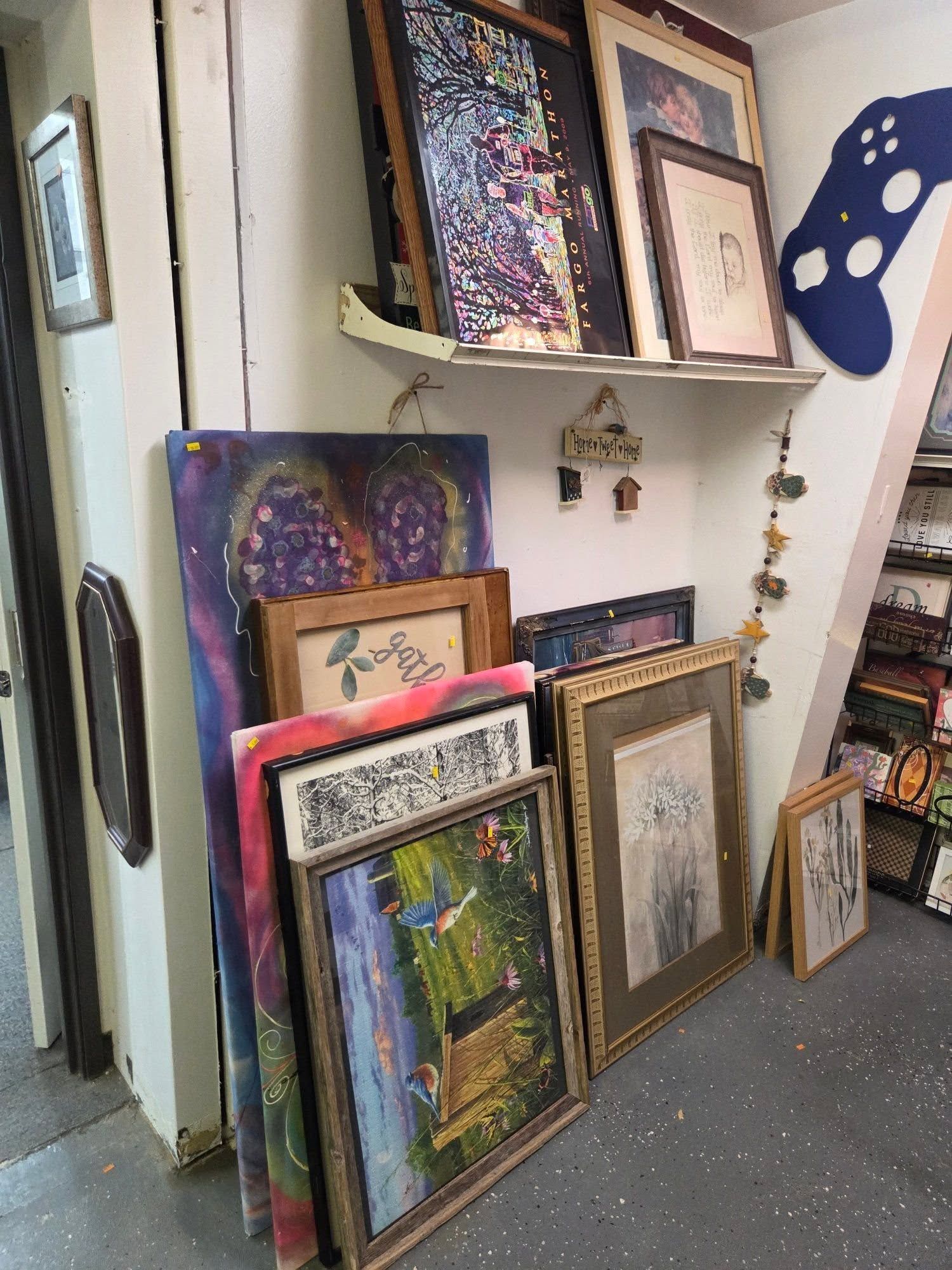 Framed artwork leaning against a wall in a gallery or studio, with colorful paintings on shelves above.