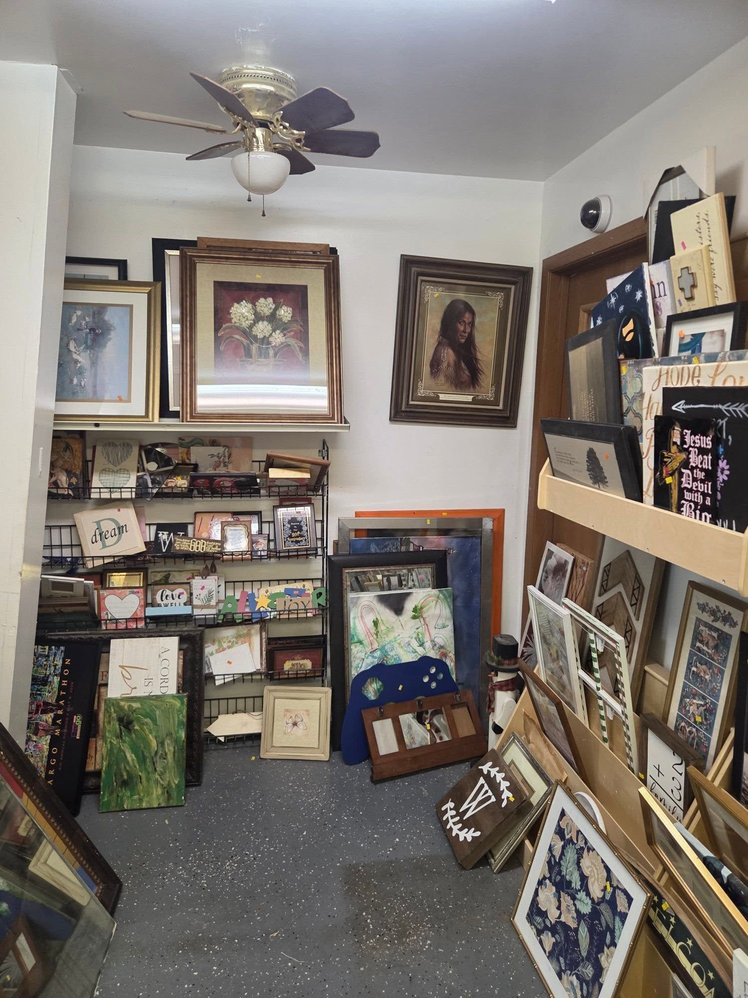 A small art-filled room with framed paintings, display shelves, and colorful quilted items on the floor.