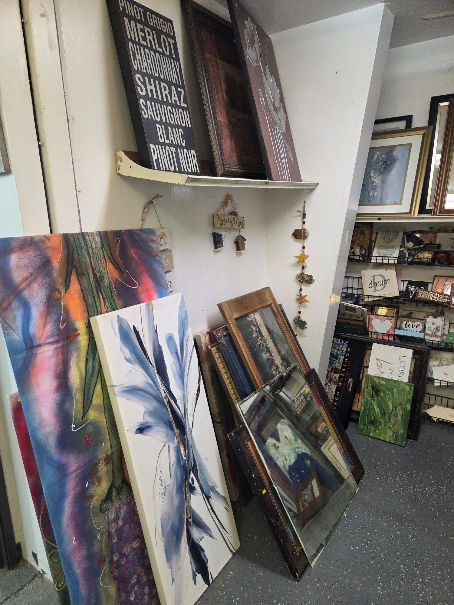 Art gallery corner with abstract paintings leaning against a wall and shelves of framed artwork.