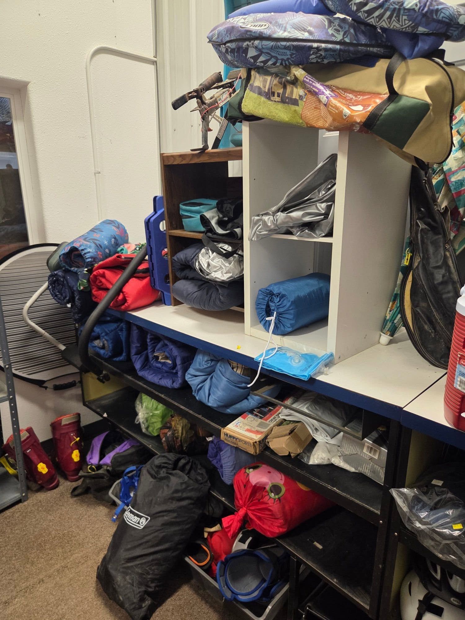 Cluttered storage shelf with stacked bags, shoes, and assorted gear in a messy room