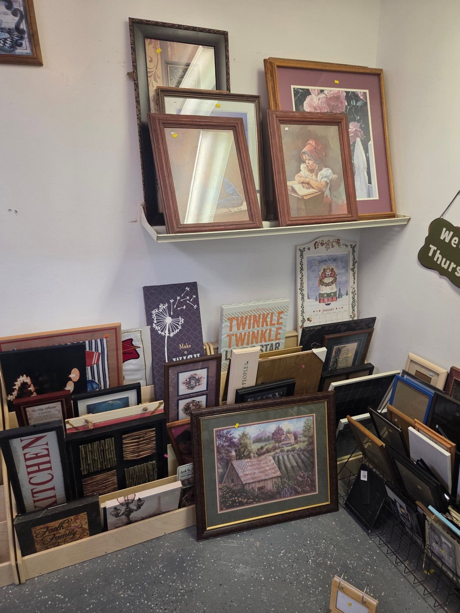 Framed artwork and photos displayed on the floor and shelf in a shop or market stall