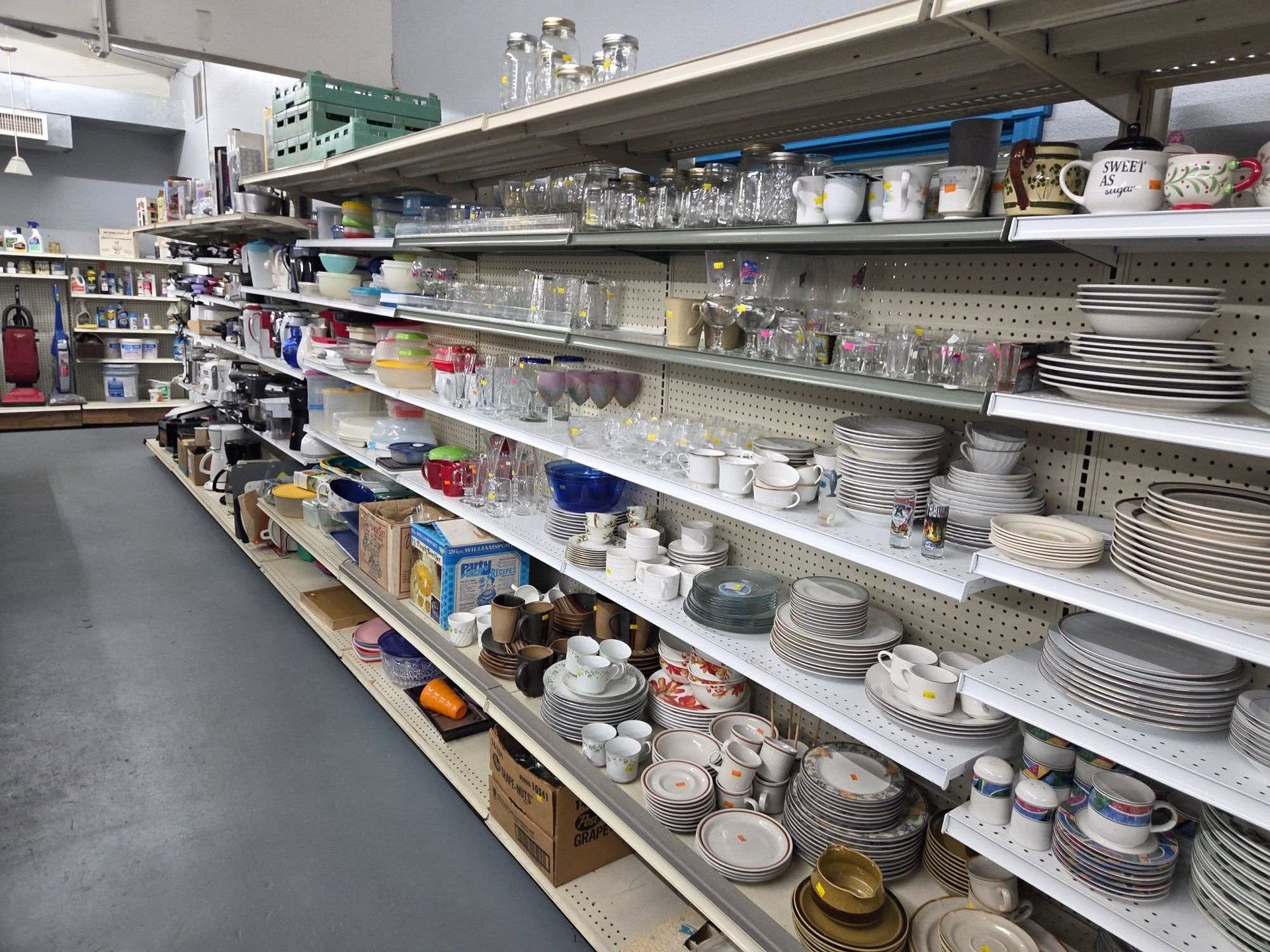 Thrift store aisle with shelves of assorted dishes, mugs, glassware, and home goods