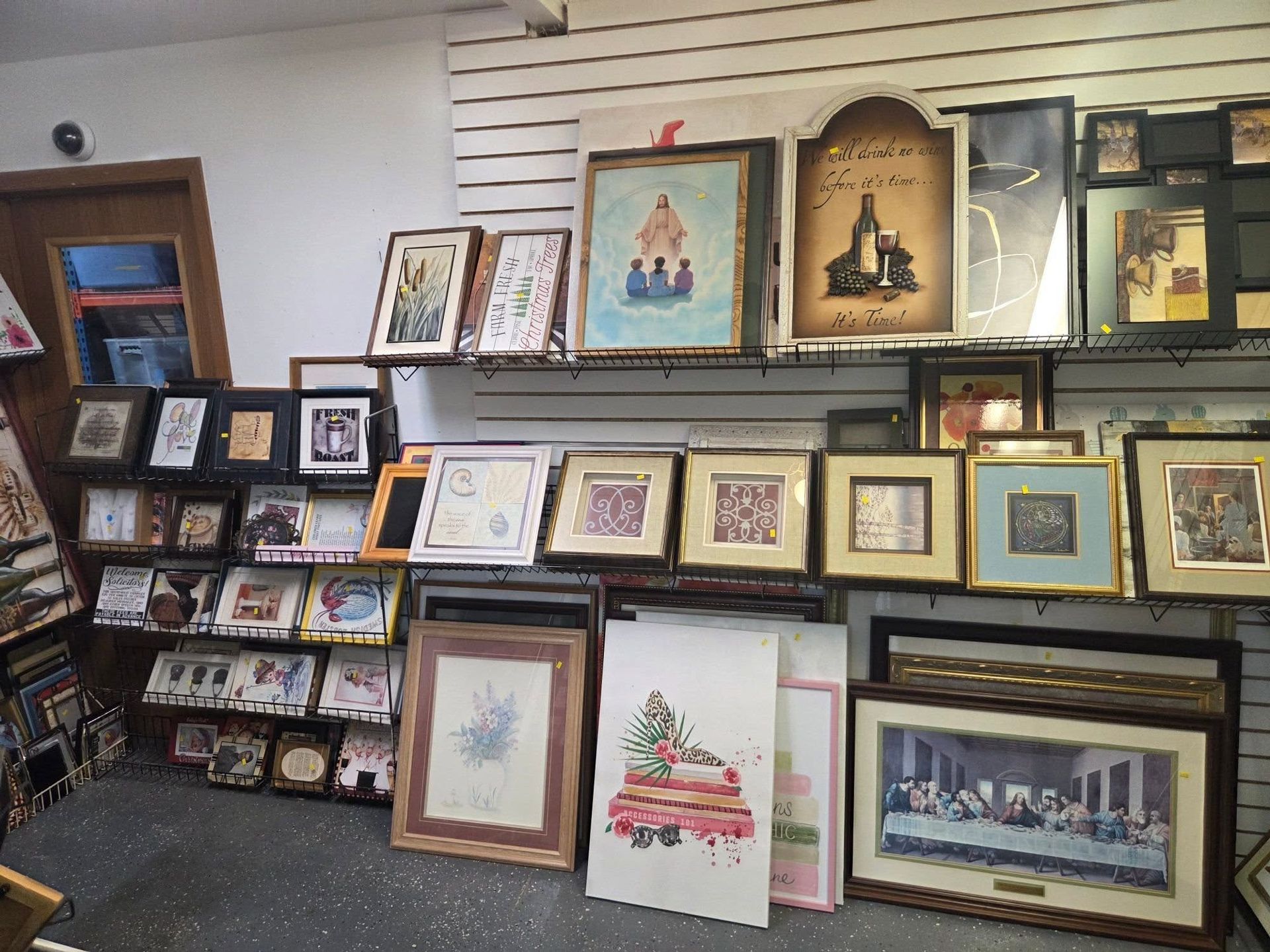 Framed art prints displayed on shelves in a shop, with colorful landscape and religious-themed pieces.
