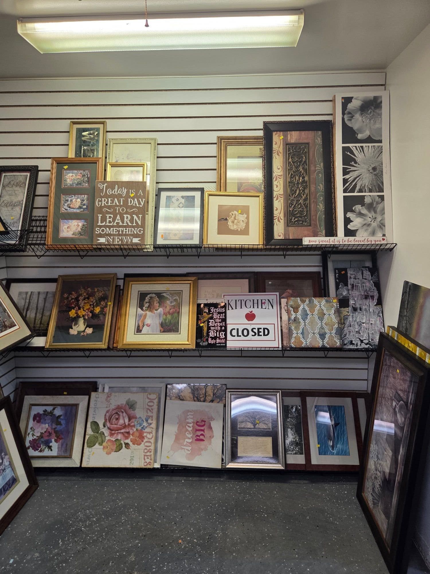 Framed art and prints displayed in a shop window, with a small “CLOSED” sign on the shelf.