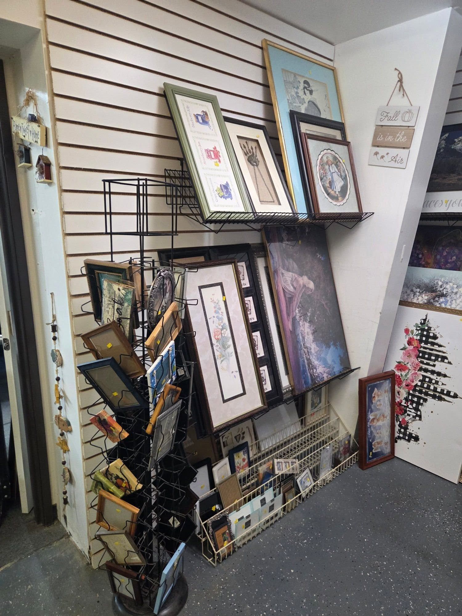 Framed artwork and prints displayed on a black wire rack in a shop aisle against white slat walls.