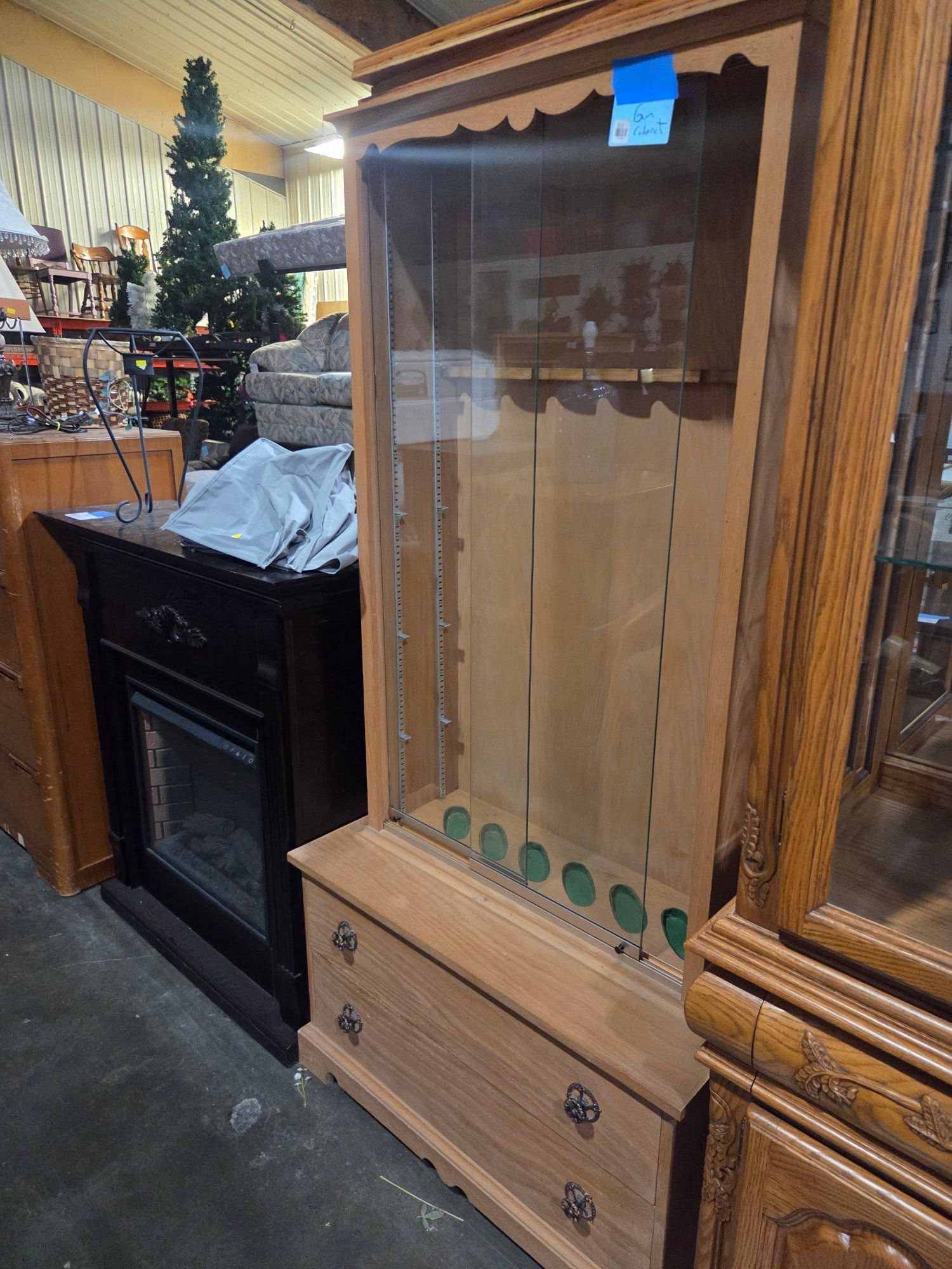 Wooden display cabinet with glass doors and drawers in a store, beside a black electric fireplace.