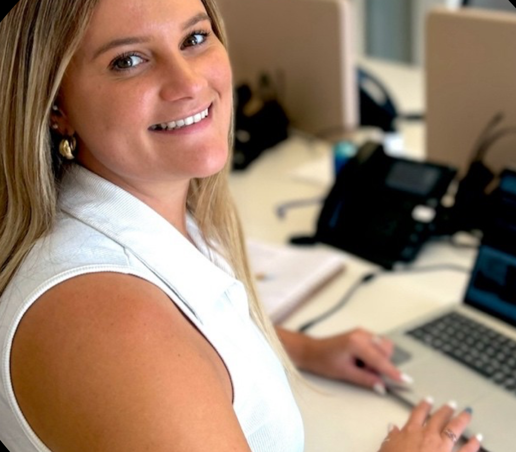 Image of Tansy Isla Adams Smit smiling at camera, working at a desk with a laptop and phone.