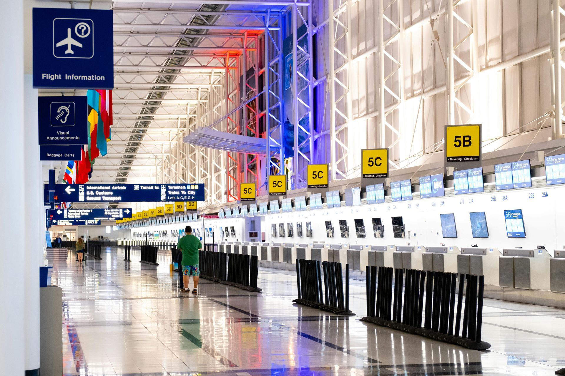 Airport check-in area with rows of kiosks, signs, and a few people walking. Blue, red, and white lighting.